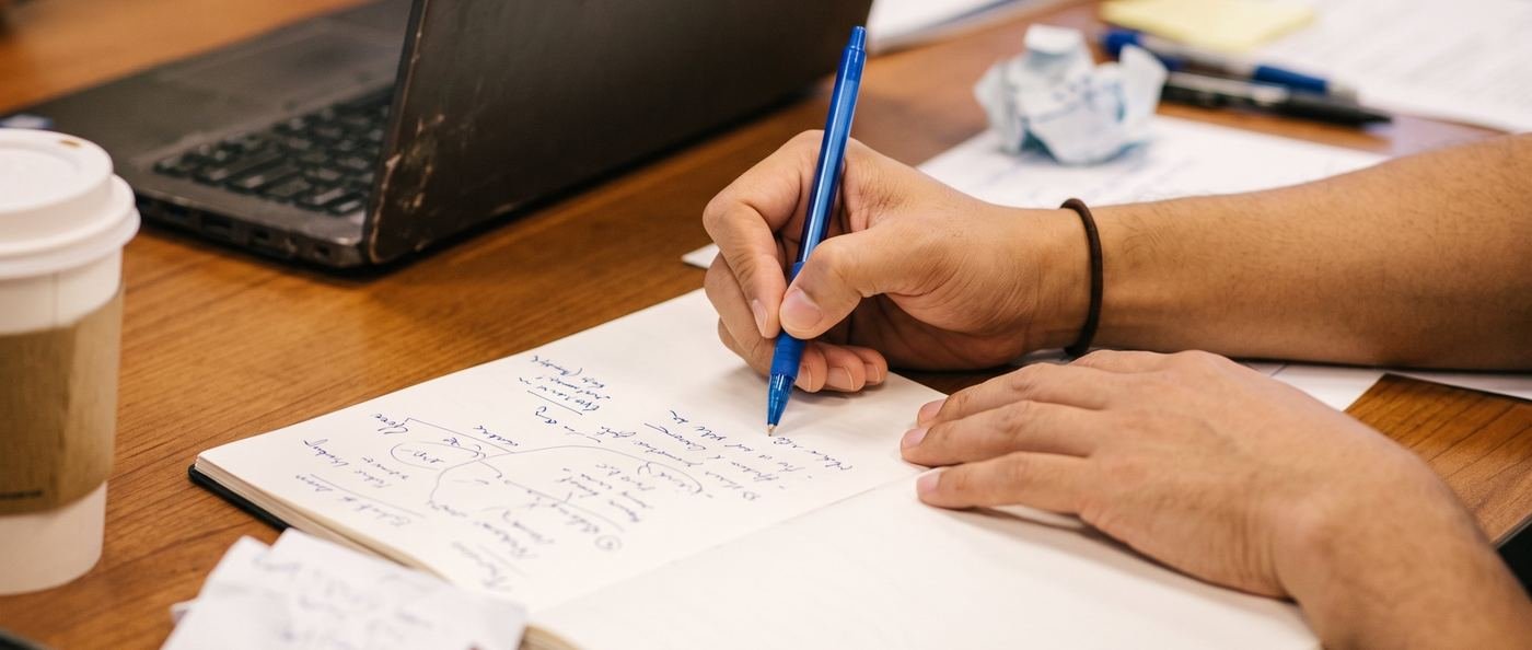 Close on two hands and a pen, working through a page of handwritten notes
    beside a laptop.